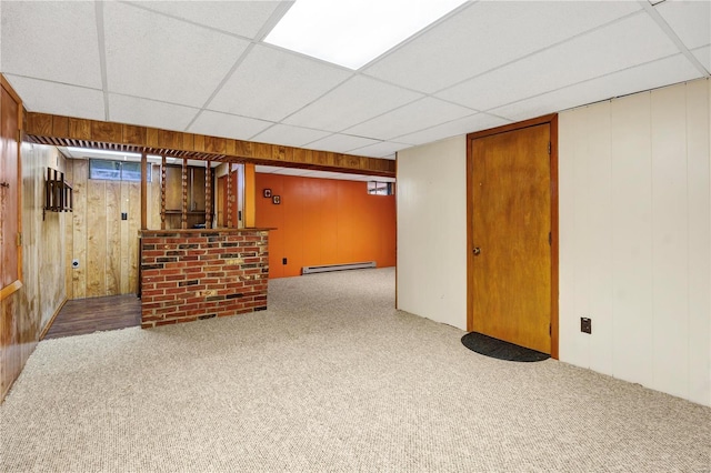 finished basement with a drop ceiling, a baseboard radiator, carpet flooring, and wooden walls