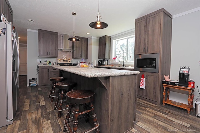 kitchen with pendant lighting, light countertops, appliances with stainless steel finishes, a kitchen island, and wall chimney exhaust hood