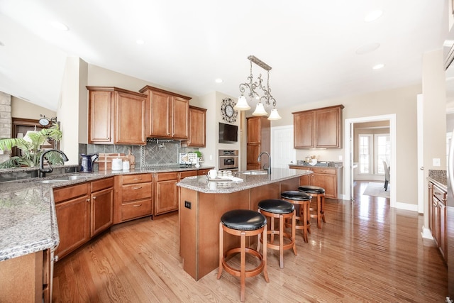 kitchen with oven, an island with sink, brown cabinetry, and a sink