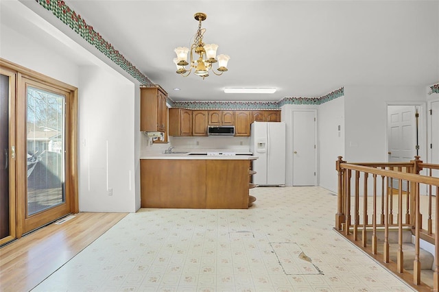 kitchen with white refrigerator with ice dispenser, brown cabinetry, stainless steel microwave, a peninsula, and a notable chandelier