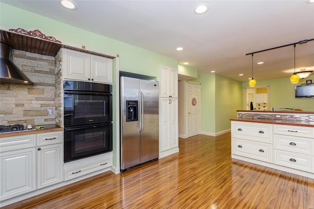 kitchen with dobule oven black, wall chimney range hood, light wood-style floors, white cabinetry, and stainless steel refrigerator with ice dispenser