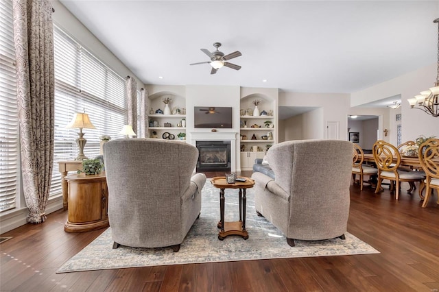 living area featuring built in shelves, visible vents, dark wood-type flooring, a glass covered fireplace, and ceiling fan with notable chandelier