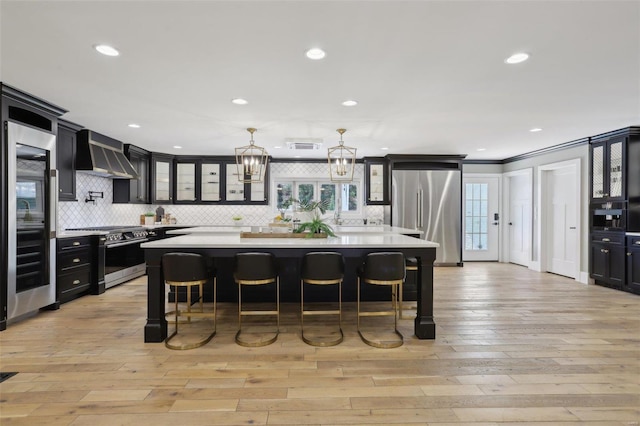 kitchen with dark cabinetry, stainless steel appliances, light countertops, a kitchen breakfast bar, and wall chimney range hood