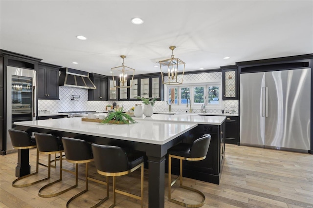 kitchen featuring light wood-type flooring, dark cabinetry, stainless steel appliances, wall chimney exhaust hood, and light countertops