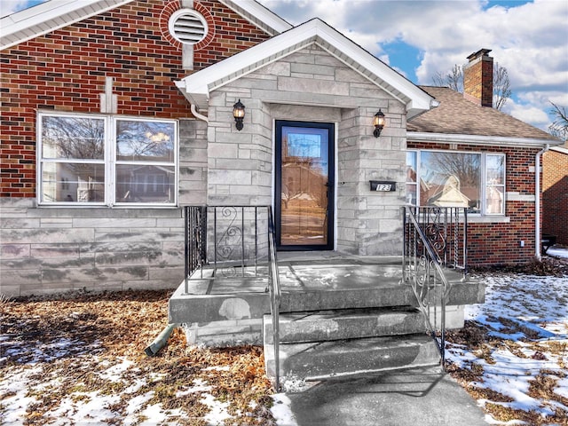 snow covered property entrance with stone siding, a shingled roof, a chimney, and brick siding