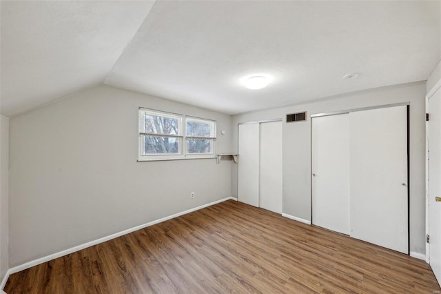 unfurnished bedroom featuring wood finished floors, visible vents, baseboards, vaulted ceiling, and multiple closets