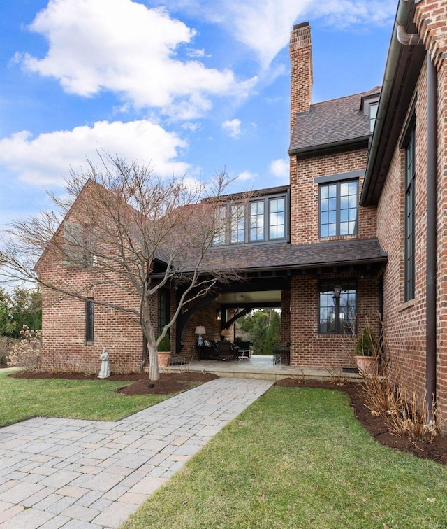 rear view of property with brick siding, a shingled roof, a lawn, a chimney, and a patio area