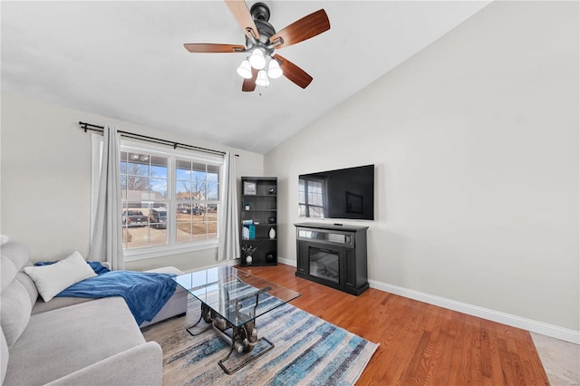 living room featuring baseboards, a glass covered fireplace, lofted ceiling, ceiling fan, and wood finished floors