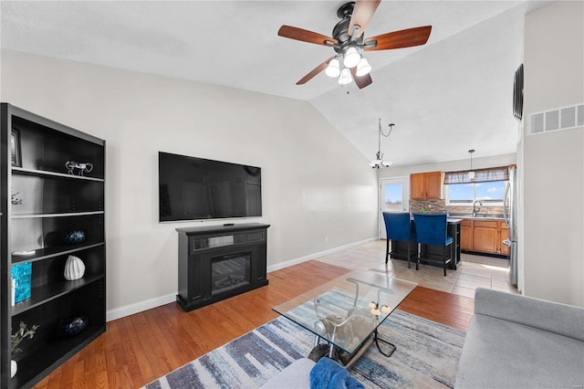 living area featuring visible vents, baseboards, a glass covered fireplace, light wood-style flooring, and vaulted ceiling