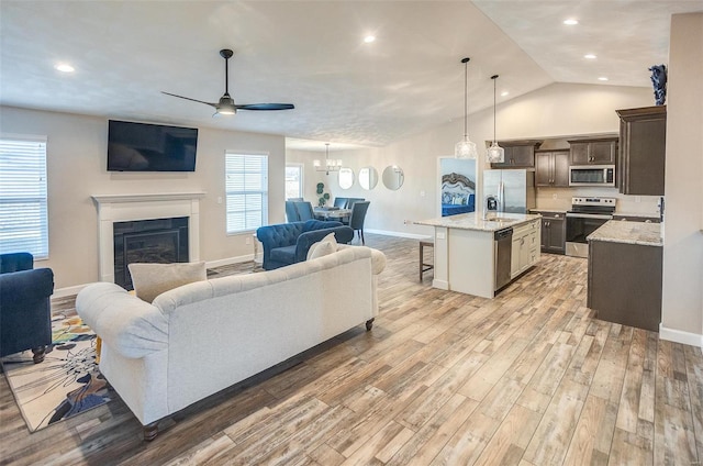 living room featuring ceiling fan with notable chandelier, a fireplace, baseboards, vaulted ceiling, and light wood-type flooring