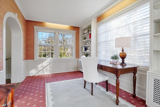 carpeted home office with built in features, arched walkways, a decorative wall, ornamental molding, and wainscoting