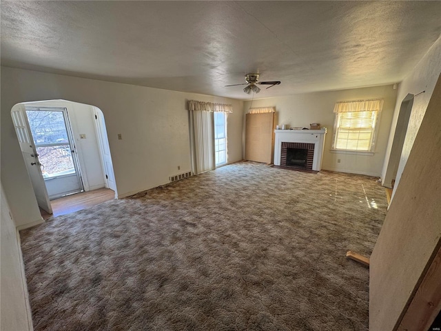 unfurnished living room with visible vents, arched walkways, ceiling fan, carpet flooring, and a brick fireplace