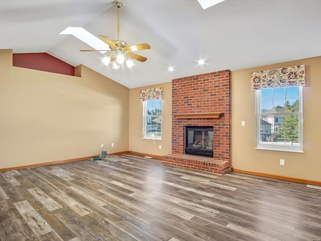 unfurnished living room with a brick fireplace, ceiling fan, wood finished floors, vaulted ceiling with skylight, and baseboards