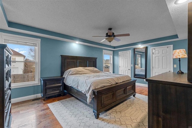 bedroom with ceiling fan, baseboards, light wood-type flooring, a tray ceiling, and a textured ceiling