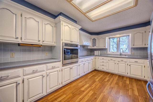 kitchen with black electric stovetop, a sink, stainless steel oven, wall chimney exhaust hood, and light wood finished floors