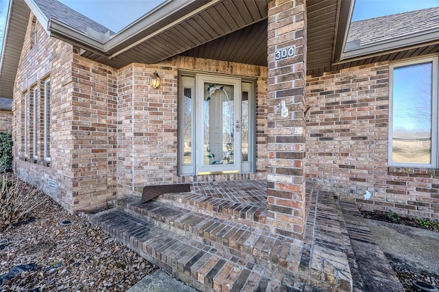 view of exterior entry with roof with shingles and brick siding