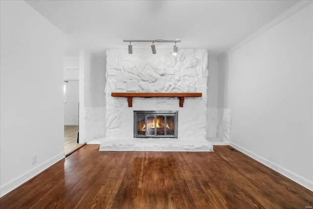 unfurnished living room featuring crown molding, rail lighting, a stone fireplace, wood finished floors, and baseboards