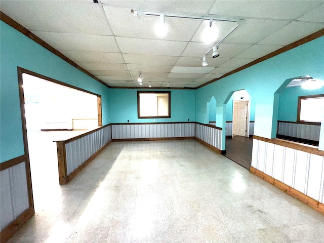 empty room with arched walkways, a wainscoted wall, a drop ceiling, and crown molding