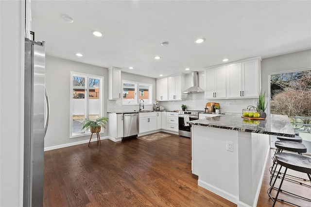 kitchen with a peninsula, stainless steel appliances, a sink, wall chimney exhaust hood, and dark wood finished floors