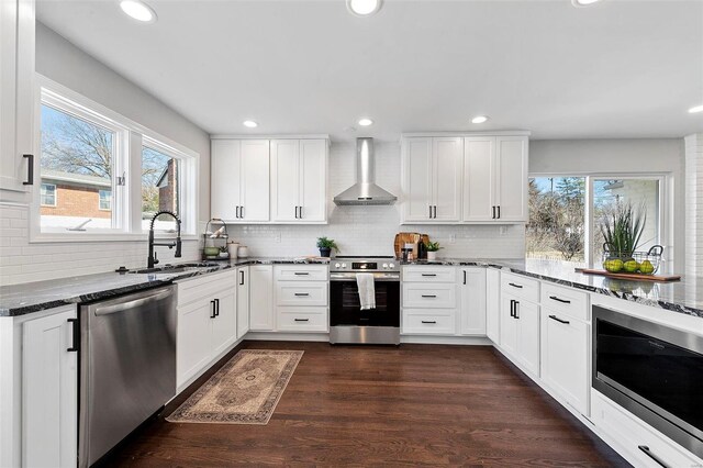 kitchen featuring appliances with stainless steel finishes, white cabinetry, a sink, dark stone counters, and wall chimney exhaust hood