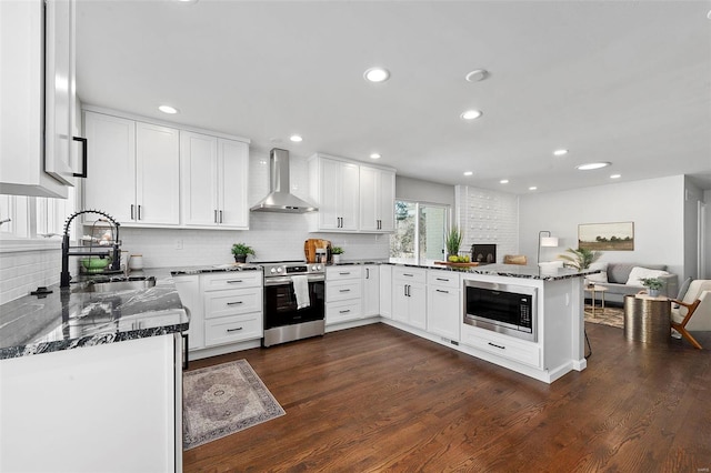 kitchen featuring stainless steel appliances, a sink, wall chimney range hood, dark stone countertops, and a peninsula