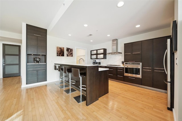 kitchen featuring light countertops, appliances with stainless steel finishes, wall chimney range hood, a kitchen bar, and a center island with sink