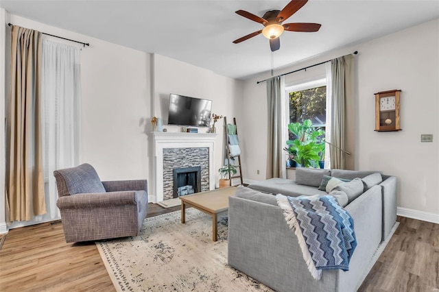 living area with baseboards, a stone fireplace, wood finished floors, and a ceiling fan