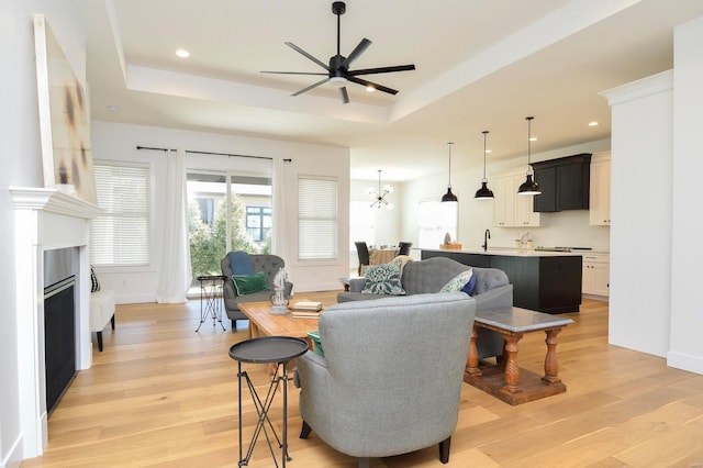 living room with light wood finished floors, a tray ceiling, ceiling fan with notable chandelier, and recessed lighting