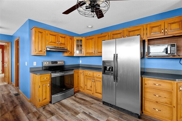 kitchen featuring dark wood-type flooring, under cabinet range hood, dark countertops, stainless steel appliances, and brown cabinetry