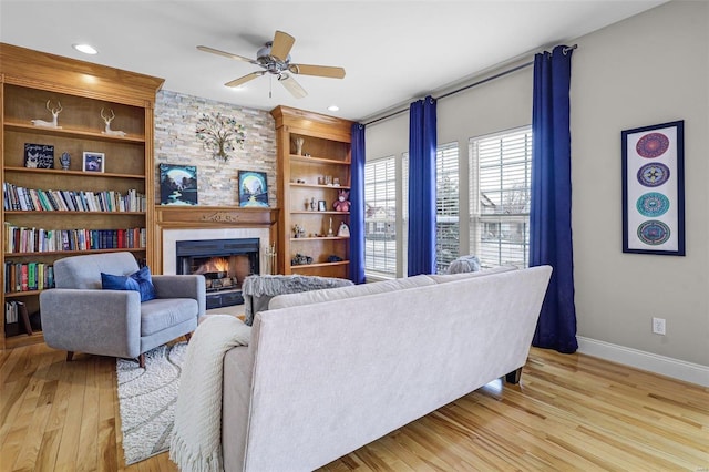 living room featuring built in features, a fireplace, ceiling fan, light wood-type flooring, and baseboards