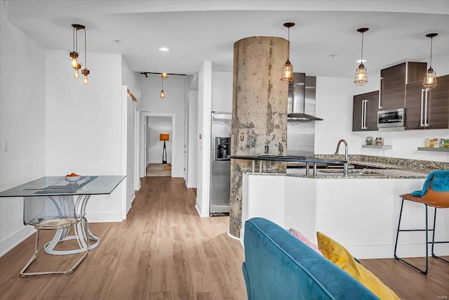 kitchen with light wood-style flooring, appliances with stainless steel finishes, a sink, dark brown cabinetry, and wall chimney exhaust hood