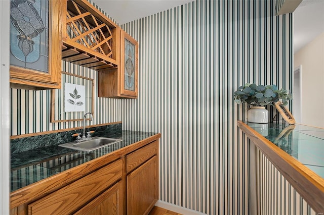 kitchen with wallpapered walls, glass insert cabinets, tile counters, brown cabinets, and a sink