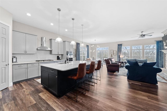 kitchen featuring dark wood-style flooring, gas stovetop, wall chimney range hood, tasteful backsplash, and an island with sink