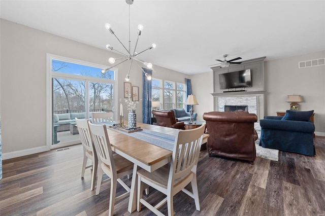 dining space with dark wood-style floors, a stone fireplace, ceiling fan with notable chandelier, and visible vents