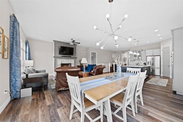 dining space featuring baseboards, ceiling fan with notable chandelier, dark wood finished floors, and a stone fireplace