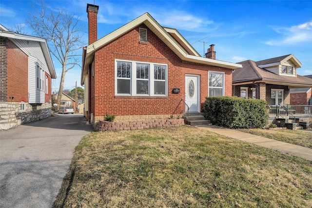 bungalow-style house featuring driveway, brick siding, a chimney, and a front lawn