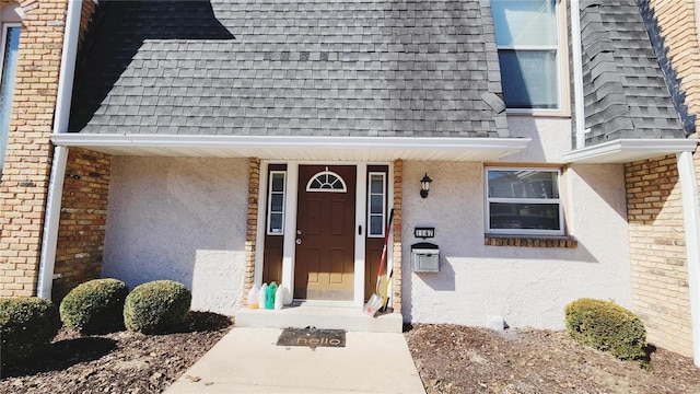 entrance to property featuring a shingled roof, mansard roof, and stucco siding