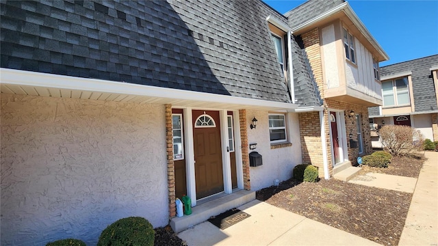 view of exterior entry featuring brick siding, stucco siding, mansard roof, and roof with shingles