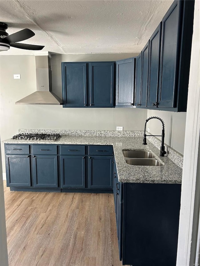 kitchen featuring stainless steel gas cooktop, blue cabinetry, light wood finished floors, a sink, and wall chimney exhaust hood