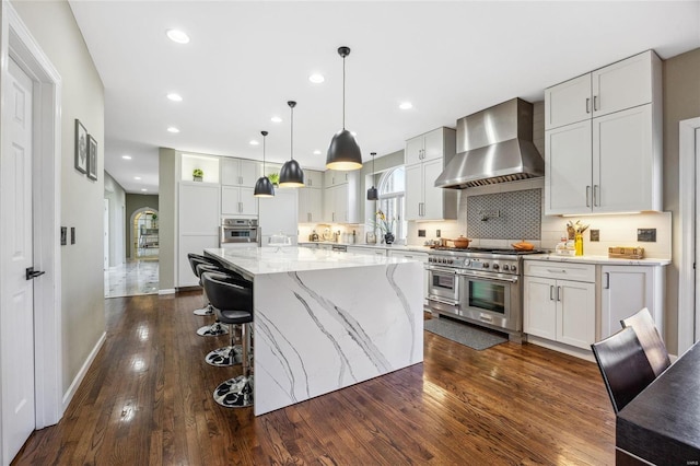 kitchen with dark wood-style flooring, backsplash, appliances with stainless steel finishes, light stone countertops, and wall chimney exhaust hood