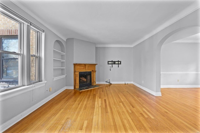 unfurnished living room featuring arched walkways, built in shelves, a fireplace, baseboards, and light wood-type flooring