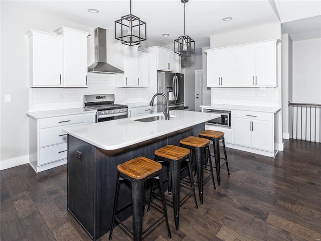 kitchen featuring a kitchen island with sink, a sink, stainless steel appliances, wall chimney range hood, and dark wood-style flooring