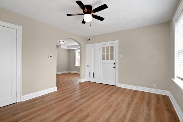 empty room featuring a ceiling fan, arched walkways, light wood-style flooring, and baseboards