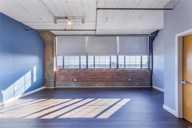 empty room featuring brick wall, wood finished floors, rail lighting, and baseboards