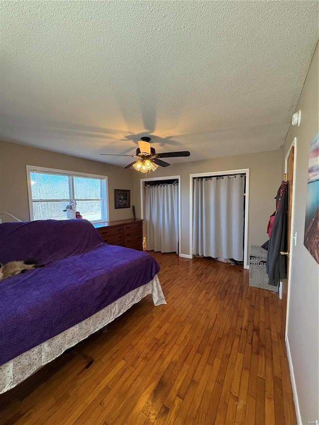 bedroom featuring ceiling fan, a textured ceiling, baseboards, hardwood / wood-style floors, and two closets