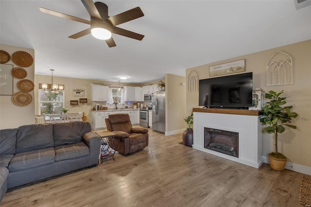 living area featuring light wood-style flooring, ceiling fan with notable chandelier, baseboards, and a glass covered fireplace