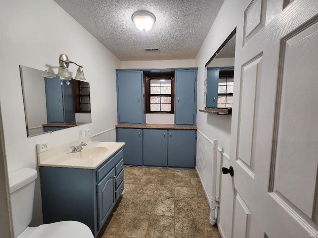 bathroom featuring a textured ceiling, toilet, visible vents, vanity, and wainscoting