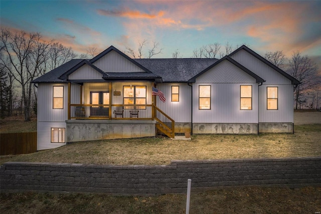 back of house at dusk with covered porch, roof with shingles, and a yard