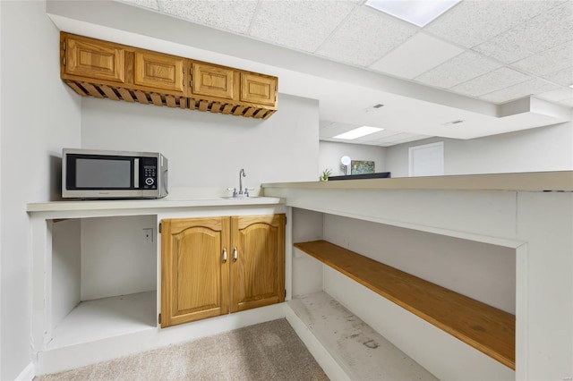 kitchen with light countertops, stainless steel microwave, brown cabinetry, a sink, and a drop ceiling