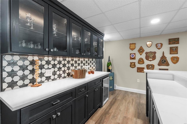 kitchen with dark cabinets, beverage cooler, wood finished floors, light countertops, and backsplash